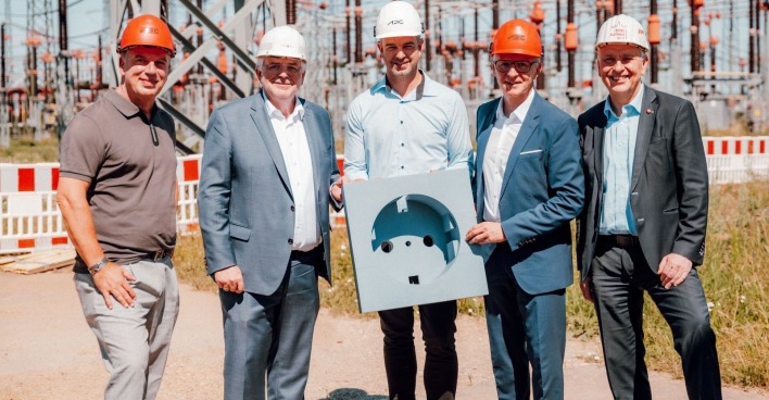 Fünf Männer in Bürokleidung mit Baustellenhelmen an einer sonnigen Baustelle
