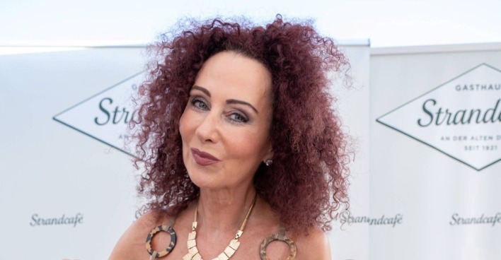 Mausi Lugner bei einem öffentlichen Auftritt in Österreich. Die Society-Lady trägt lockiges Haar, eine goldene Statement-Kette und lächelt in die Kamera.