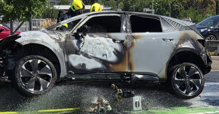 Fahrzeugbrand: Ausgebranntes E-Auto nach Fahrzeugbrand beim Bahnhof Kapfenberg mit Feuerwehrleuten im Hintergrund.