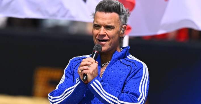 Robbie Williams bei einem Auftritt auf der Bühne im blauen Trainingsanzug mit Mikrofon. Er spricht über seine Autismus-Diagnose