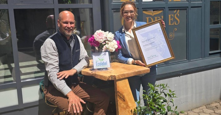 Die Besitzer des Villacher Boutique Hotel Goldenes Lamm, Jakob und Nicole Ottacher, die auf einer Bank vor ihrem Betrieb sitzen und eine Urkunde für die Zertifizierung mit dem EU Ecolabel in die Höhe halten
