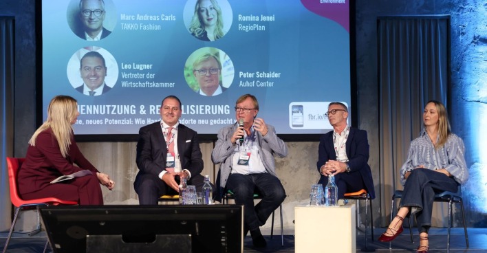 Drei Männer und zwei Frauen sitzen Vor einer Videoleinwand am Podium und diskutieren