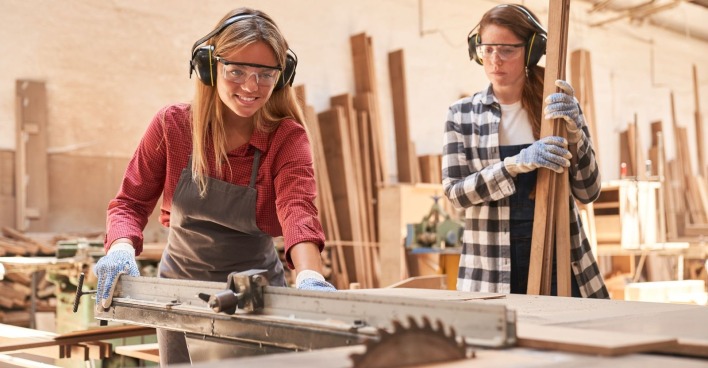 Zwei junge Damen - eine mit langem blonden Haaren, eine mit langem dunklem Haar - arbeiten an einer Kreissäge. Beide tragen Schutzbrille und Arbeitskleidung. Eine hält Holzlatten, die andere schneidet ein Brett.