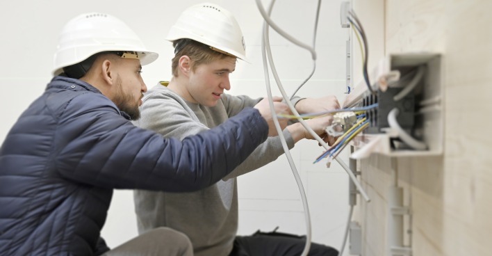 Zwei junge Personen mit weißen Schutzhelmen arbeiten gemeinsam mit Baumaterialien.