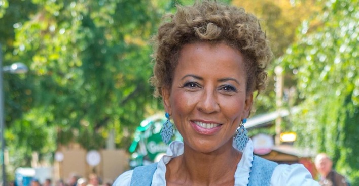 Arabella Kiesbauer lächelt in die Kamera. Die österreichische Moderatorin trägt ein blau-weißes Outfit und auffällige blaue Ohrringe.