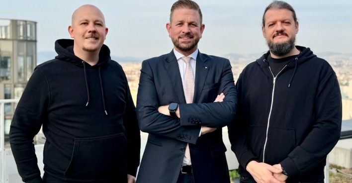 Drei Männer in Anzug beziehungsweise dunklen Hoodies stehen auf einem Balkon