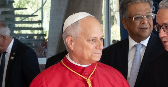 Kirchenvertreter in rotem Gewand mit weißem Scheitelkäppchen im Gespräch mit mehreren Männern bei einem öffentlichen Termin.
