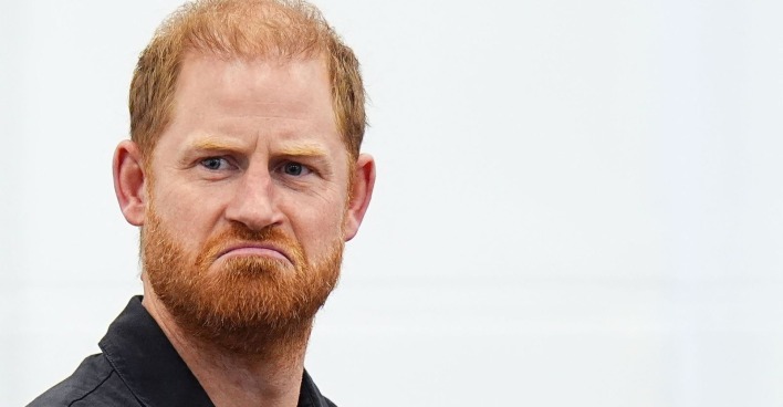 Nahaufnahme von Prinz Harry mit ernstem Gesichtsausdruck, rotem Bart und schwarzem Oberteil vor hellem Hintergrund.