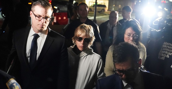 Selma Blair mit Sonnenbrille und grauem Kapuzenpullover, begleitet von einem Mann im Anzug und mehreren Einsatzkräften bei Nacht.