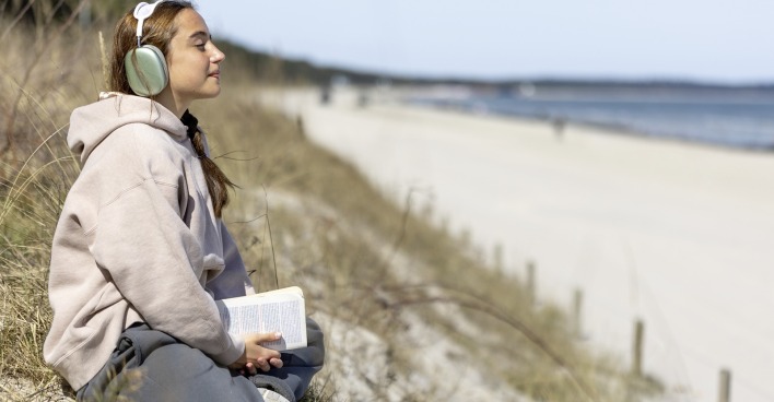 Weiblicher Teenager sitzt mit einem Buch in der Hand und mit aufgesetztem Kopfhörer am Strand
