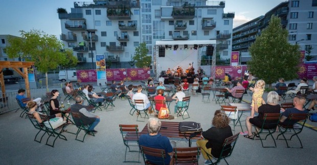 Grüpchen von Menschen sitzend, Holztische, eine Bühne und Band auf einem einem offenen Platz. Häuser im Hintergrund, früher Abend.