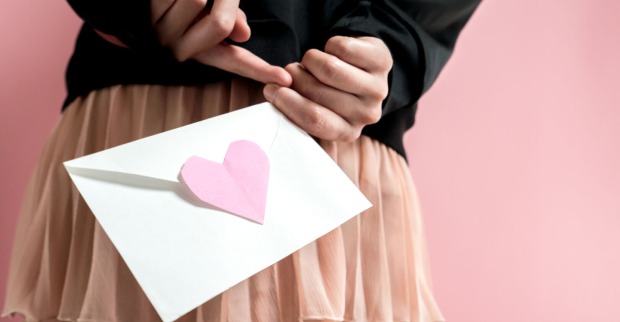 Ein Mädchen hält einen Brief mit einem rosa Herz hinter dem Rücken. Man sieht nur ihre Hände, den Brief und ihren Rock.