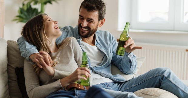 Ein Mann und eine Frau sitzen lachend Arm in Arm mit Bier auf einer Couch