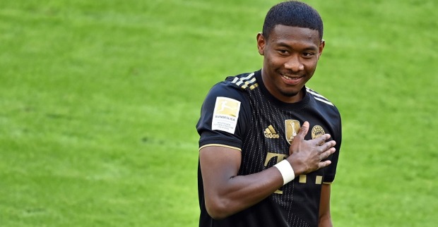 David Alaba hält seine Hand auf das Klublogo seines schwarzen Bayern München Dress und lacht.