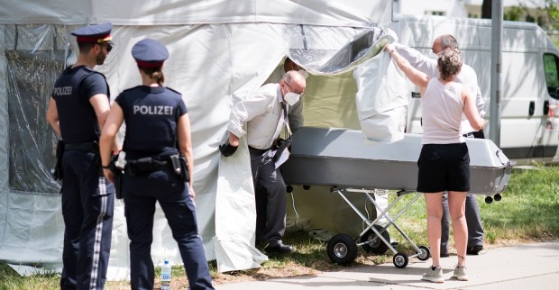 Zwei Polizisten bewachen ein Obduktionszelt. Eine Frau hält den Vorhang zur Seite. Zwei Männer mit Mundschutz schieben einen Blechsarg nach draußen.