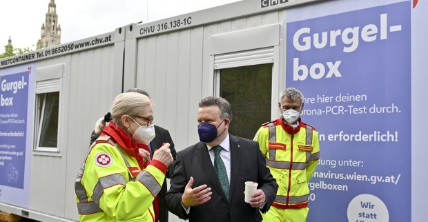 Bürgermeister Michael trägt eine Maske. Er steht mit zwei Sanitätern vor einer Gurgelbox.