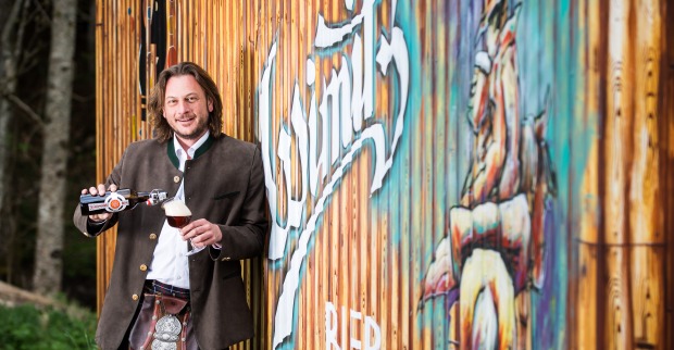 Josef Habich steht im Kärntner Kilt vor dem Logo seiner Brauerei und schenkt sich ein dunkles Bier ein