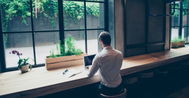 Mann von hinten, der in einem Café vor seinem Notebook sitzt - mit Blick aus dem Fenster auf einen begrünten Innenhof | Credit: iStock.com/Deagreez
