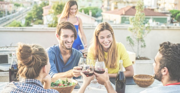 Junge Menschen genießen eine gemeinsame Mahlzeit unter freiem Himmel auf einer Dachterrasse | Credit: iStock.com/DisobeyArt