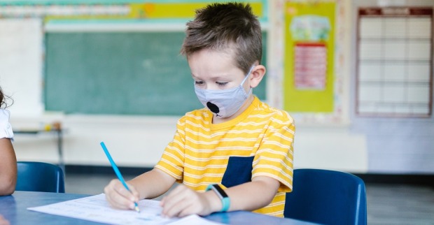 Ein Bub sitzt in einem Klassenzimmer und schreibt. Er trägt einen Mundnasenschutz.