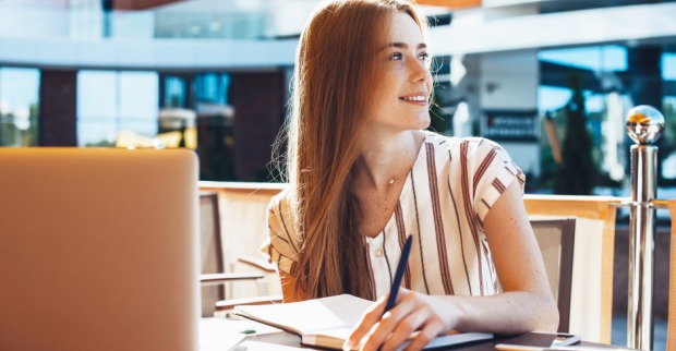 Rothaarige Studentin an einem sonnigen Tag in einem Straßencafé, den Notebook vor sich aufgeklappt | Credit: iStock.coM/Strelciuc Dumitru