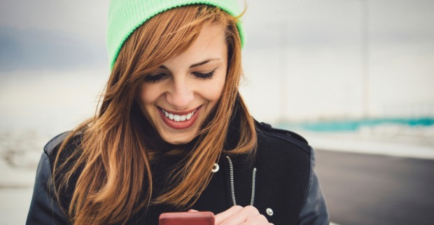 Glückliche Frau blickt während einem Spaziergang an einem kalten Wintertag auf ihr Smartphone | Credit: iStock.com/DjordjeDjurdjevic