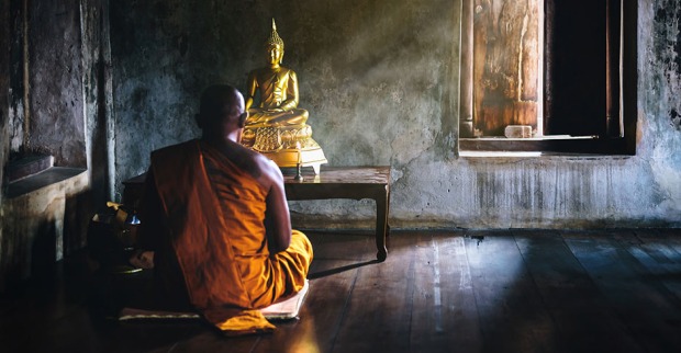 Buddhistischer Mönch in Thailand sitzt im Schneidersitz vor einer Buddha-Statue | Credit: iStock.com/flukyfluky