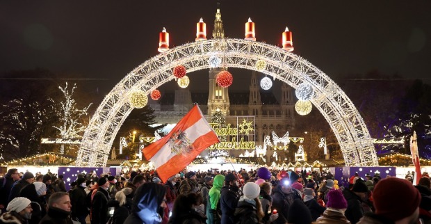 Menschen mit Fahnen demonstrieren vor dem Wiener Christkindlmarkt. Im Hintergrund Weihanchtsbeleuchtung