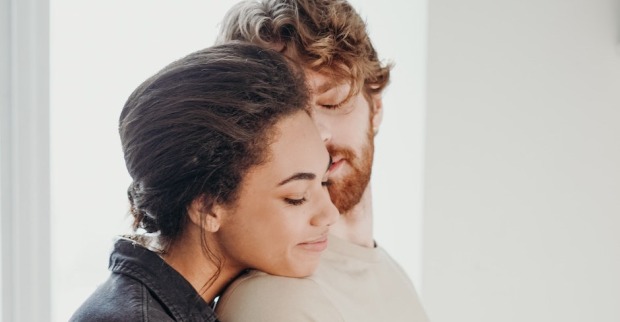 Eine Frau umarmt ihren Partner von hinten. Beide haben glücklich die Augen geschlossen