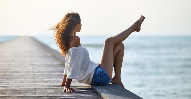 Junge Frau in Jeansshorts sitzt auf einem Meeressteg und genießt die Aussicht | Credit: iStock.com/Viktor_Gladkov