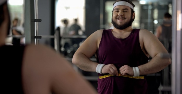 Ein übergewichtiger Mann steht vor einem Spiegel im Fitnessstudio und misst seinen Bauchumfang mit einem Maßband