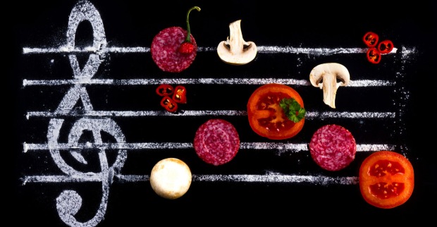 Eine schwarze Tafel, auf der mit Kreide ein Notenschlüssel und fünf Taktstriche aufgemalt wurden, auf denen Pilze, Tomaten und Salamischeiben platziert wurden