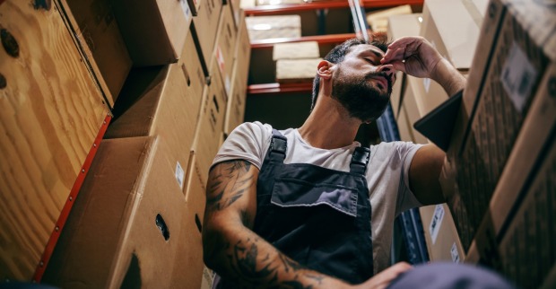 Erschöpfter, bärtiger Mann in einem Lagerraum. | Credit: iStock.com/dusanpetkovic
