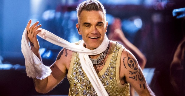 Robbie Williams in einem goldglitzernden Oberteil und einem weißen Schal bei einem Auftritt.