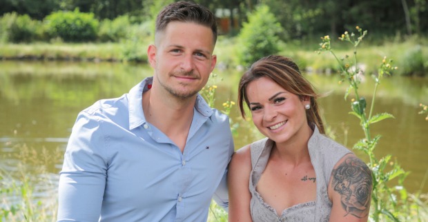 Stefan mit einem hellblauen Hemd und Nicole mit einer Tätowierung am Arm sitzen vor einem See.