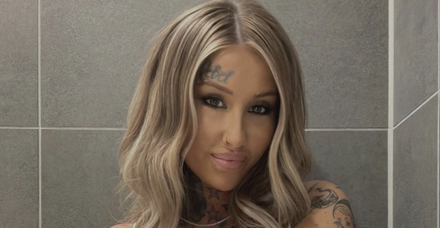 Blonde Frau mit Tattoo im Gesicht vor einer Fliesenwand
