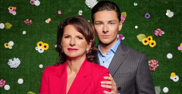 Claudia Obert in einer roten Bluse mit ihrem Partner Max vor einer grünen Wand mit bunten Blumen.