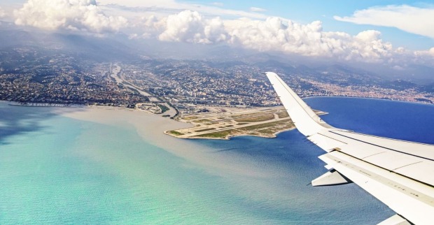 Die Tragfläche eines Flugzeugs aus dem fliegenden Flugzeug fotografiert. Darunter türkises Meer und eine Landebahn