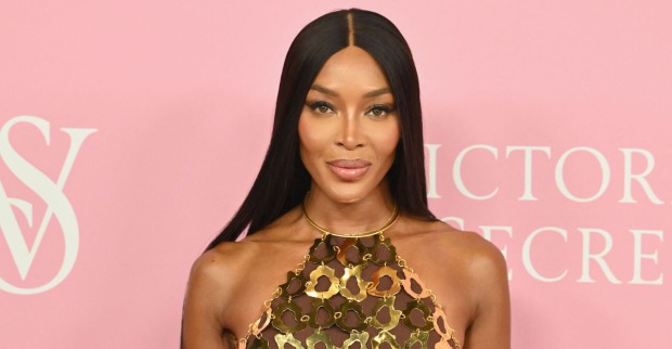 Naomi Campbell mit langen schwarzen Haaren und einem goldenen Kleid vor einer rosa Victoria Secrets Wand.