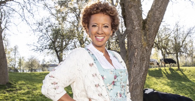 Arabella Kiesbauer in einem hellblauen Dirndl mit einer weißen Weste auf einer Kuhwiese.