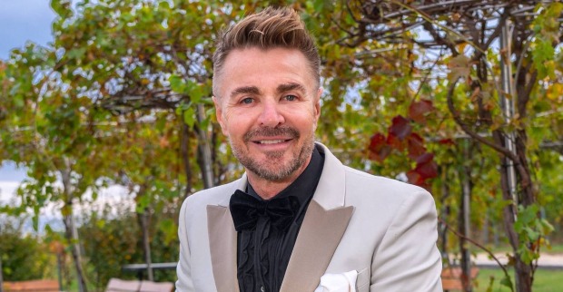 Leo Hillinger mit aufgestellten blonden Haaren und einem grauen Sakko vor einer Weinrebe.