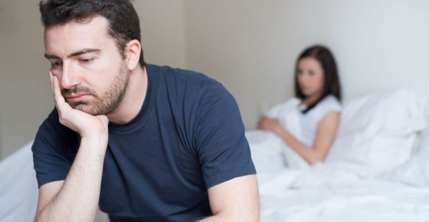 Ein Mann sitzt an den Bettkante, den Kopf in die Hand gestützt. Er wirkt nachdenklich bis bedrückt. Im Bett liegt eine Frau.