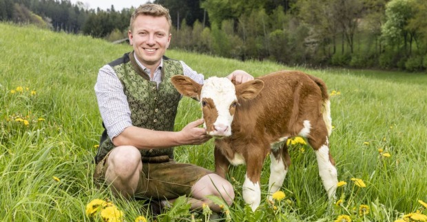 Bauer Johannes kniet auf einer Weide und streichelt ein Kalb. Er trägt eine grüne Tracht und braune Lederhosen.