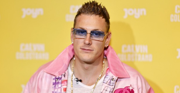 Der Reality Tv Star Calvin Kleinen trägt eine blaue Brille und eine pinke Jacke. Er steht vor einer gelben Fotowand.