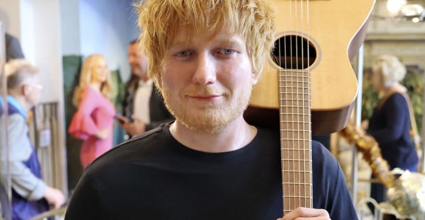 Ed Sheeran trägt ein schwarzes T-Shirt und hält eine Gitarre auf den Schultern.