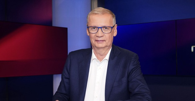 Günther Jauch trägt einen dunkelblauen Anzug und eine weiße Bluse. Er sitzt vor einem blau-roten Hintergrund.