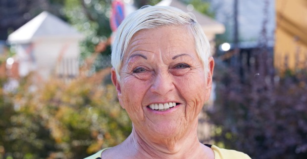 Jazz Gitti steht vor einer Wiese und Häusern. Sie trägt kurze platinblonde Haare und lächelt in die Kamera.
