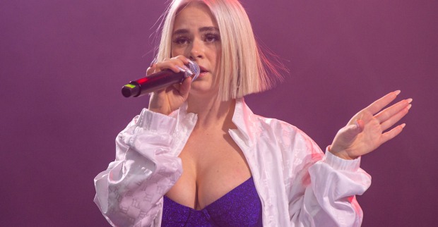 Mathea trägt ein glitzernes Bikinitop und ein weißes Hemd. Sie hat einen blonden Longbob und singt ein Lied.