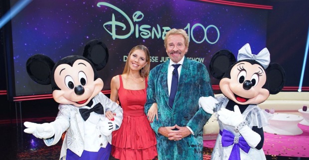 Thomas Gottschalk trägt einen blauen Anzug. Victoria Swarovski ein rotes, kurzes Kleid. Neben ihnen stehen Mickey und Mini Maus.