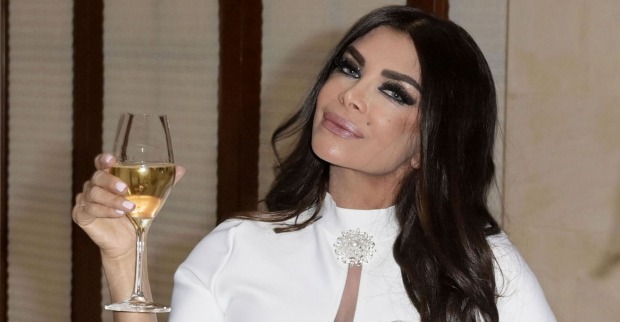 Kader Loth mit einem weißen Kleid und ein Glas Sekt in der Hand. Sie hält den Kopf schief in die Kamera und hat stark geschminkte Augen, sowie lange schwarze Haare.